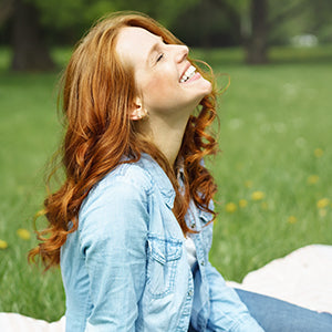 A woman with long hair sitting in an open field surrounded by trees while looking up and smiling