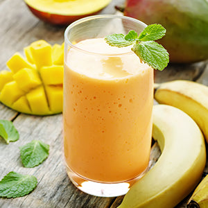 An orange smoothie in a glass over a wooden table with a mango and banana in the background.