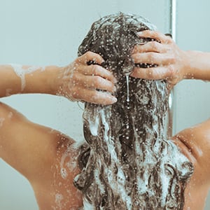 A woman is applying shampoo in the shower. Selenium sulfide is commonly found in cream, foam, shampoo, or in liquid form.